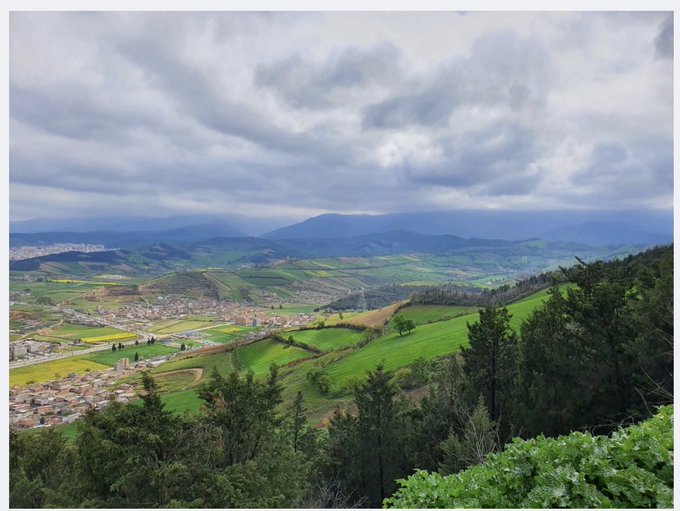 A scenic landscape view shows rolling green hills, a patchwork of yellow and green fields, scattered villages, and mountains in the distance beneath a cloudy sky.