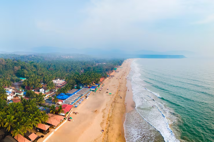 An aerial view of a tropical coastline featuring a long sandy beach, lush palm trees, beachside buildings with red and blue roofs, and gentle ocean waves.