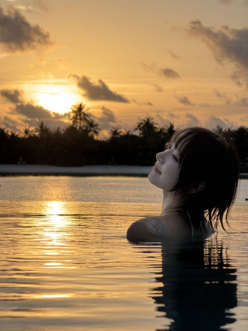 A woman with wet hair relaxes in calm waters, her face tilted upward toward the sky as a golden sunset reflects across the surface, framed by silhouetted palm trees on a distant island. The image perfectly captures the tranquil end of the rainy season at Patina, with the setting sun burning itself out over the horizon as described in the post.