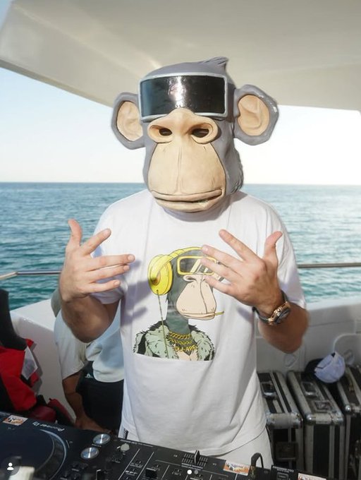 A DJ wearing a large monkey mask and a white t-shirt featuring a Bored Ape Yacht Club graphic, standing behind Pioneer DJ equipment on a boat.