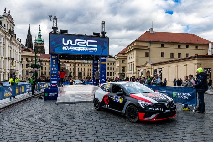 A Hyundai rally car emblazoned with "HIVE" and "WEB3 IS" livery drives onto the WRC-branded starting ramp in a cobblestone plaza in Prague for the Central European Rally, with sponsor banners for "Skupina ČEZ" and "Ševen" visible around the ceremonial start.
