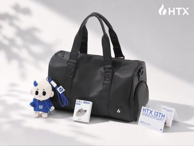 A black HTX 13th Anniversary duffel bag displayed alongside a small plush mascot, with promotional materials reading 'HTX 13TH ANNIVERSARY' and Chinese text celebrating the company.