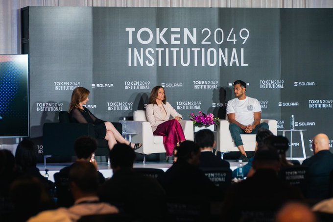 Three people sit on white armchairs on stage at the TOKEN2049 Institutional conference, with a dark backdrop displaying the event's branding and Solana logos, while an audience watches from the foreground.