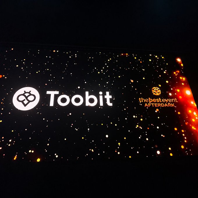 A sponsor backdrop at the AFTERDARK event in Singapore displays the "Toobit" logo and name alongside the tagline "thebestevent AFTERDARK," set against a dark, bokeh-lit background. The image visually represents Toobit's sponsorship presence at the event mentioned in the post, highlighting the company's role in supporting the evening's gathering.