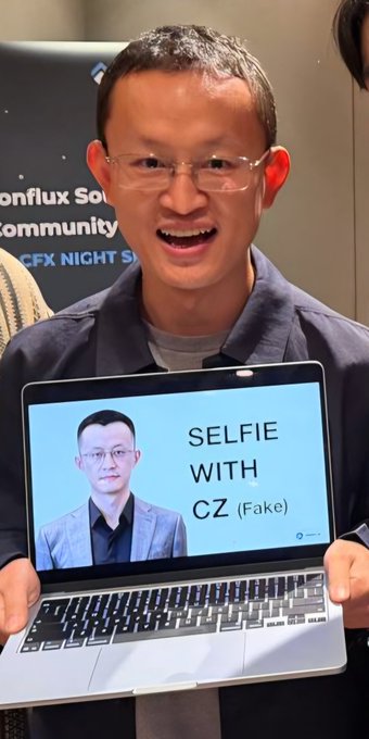 A man smiling while holding a laptop displaying an image labeled "SELFIE WITH CZ (Fake)", with "Bonflux" and "CFX NIGHT S..." visible in the background.