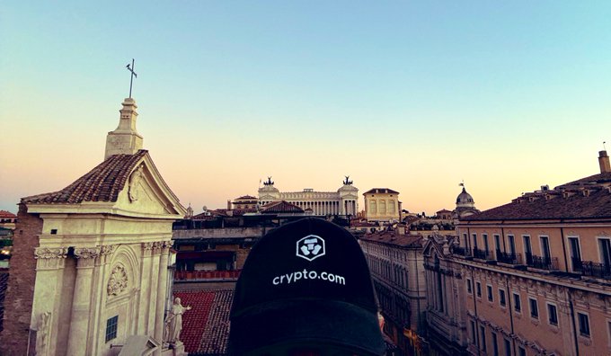 A view of a cityscape at sunset featuring historic European buildings and domes, with a black cap in the foreground displaying the 'crypto.com' logo.