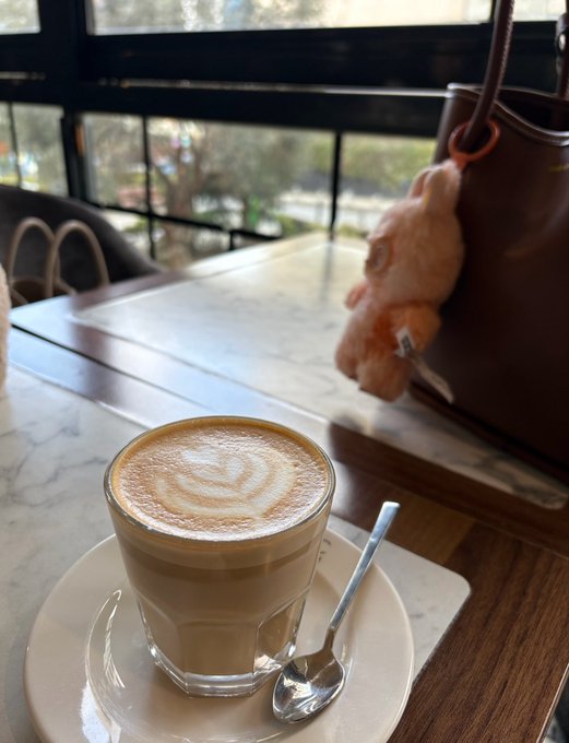 A latte with latte art sits on a marble table beside a handbag adorned with a teddy bear charm, captured in a cozy café setting. The morning coffee scene pairs nicely with the upbeat crypto greeting, suggesting a relaxed start to a bullish day in the markets.
