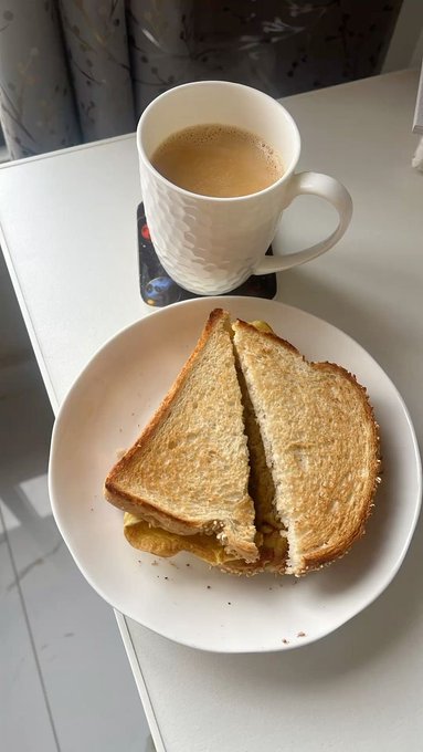 A simple breakfast of a toasted sandwich cut diagonally on a white plate, accompanied by a white mug of coffee with milk, sitting on a white table.