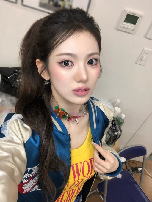 A young woman with long dark hair poses in a makeup room, wearing a blue and cream bomber jacket over a yellow shirt with partial text reading 'WO' and 'CR', along with a colorful spiked necklace and star earrings.