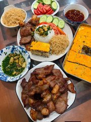 An overhead view of a dining table laden with various dishes, including a plate of glazed meat, yellow crepes or omelets, sliced cucumbers, and a small bowl of dipping sauce.