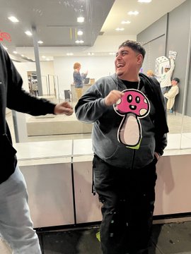 A man in a gray mushroom sweatshirt and black pants smiles while standing in what appears to be a brightly lit store or food court.