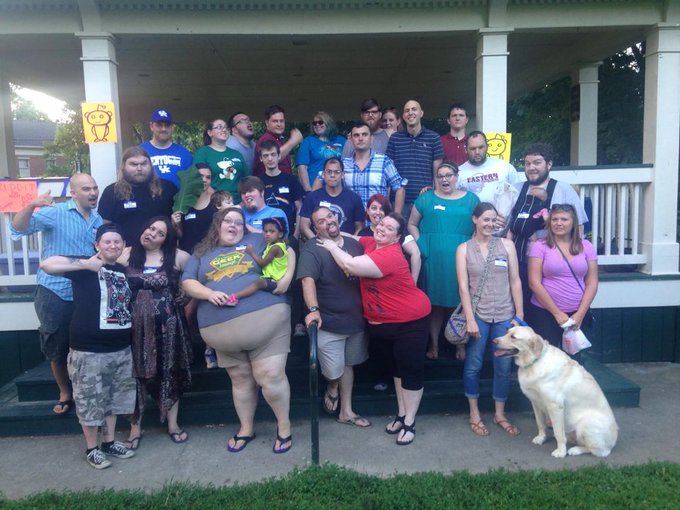 A diverse group of people poses together on the steps of a house with a "BitHouse" sign, alongside a yellow Labrador, celebrating a successful local Bitcoin meetup. One person wears a "Western Kentucky" shirt, and another sports a green t-shirt with the Bitcoin logo, highlighting the community's enthusiasm for cryptocurrency.