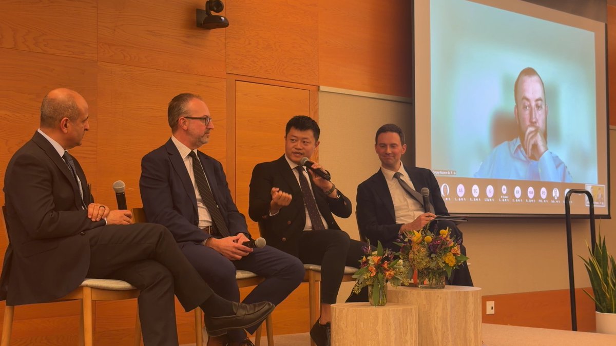 A panel discussion with four men in business attire seated on stage, with one man speaking into a microphone while a video call participant is displayed on a large screen behind them.