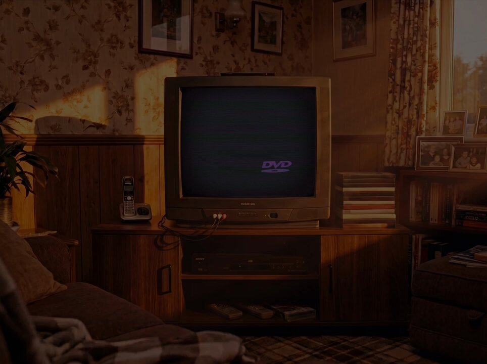 The image shows a living room with an old television set sitting on top of a wooden entertainment center, which also houses other electronics such as a DVD player and a phone. The TV appears to be turned off or muted, indicating that no one is watching it at the moment. In front of the television, there are two couches - one placed against the wall and another located in the middle of the room. A potted plant can also be seen on the left side of the image.
