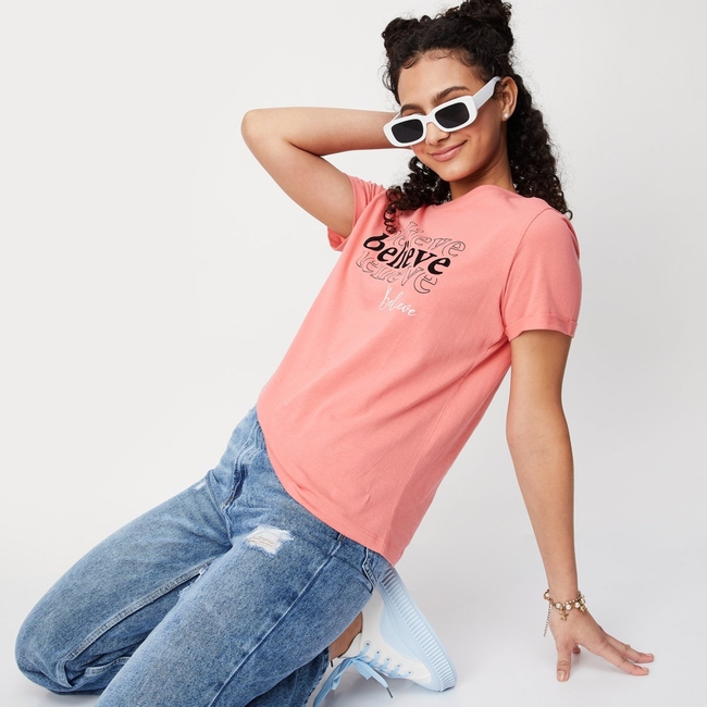 A woman with curly hair kneels while wearing white sunglasses, a coral pink t-shirt with "Believe" text, distressed blue jeans, and white sneakers.

Note: The accompanying post contains spam promoting gambling, betting, and hash game websites, which appears unrelated to the image content of a model wearing casual clothing.