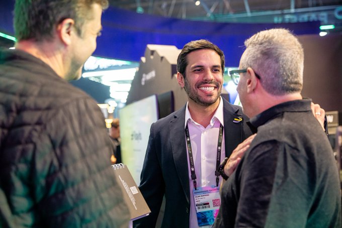 Three men conversing at what appears to be a conference or trade show, with one wearing a lanyard and smiling, and Hebrew text visible in the background.