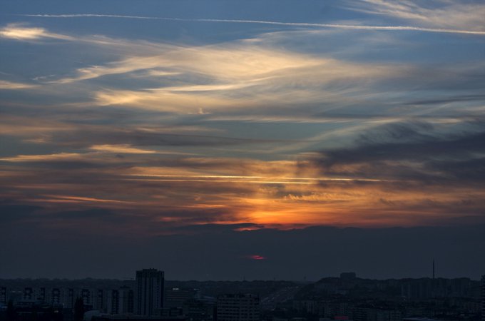 A sunset sky filled with dramatic, layered clouds hangs over the silhouette of a distant cityscape.