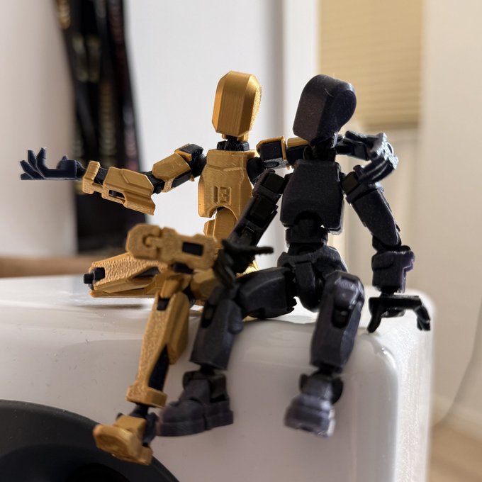 Two small articulated robot figurines, one gold and one black, posed sitting on the edge of a white surface, resembling miniature action figures or 3D-printed toys.