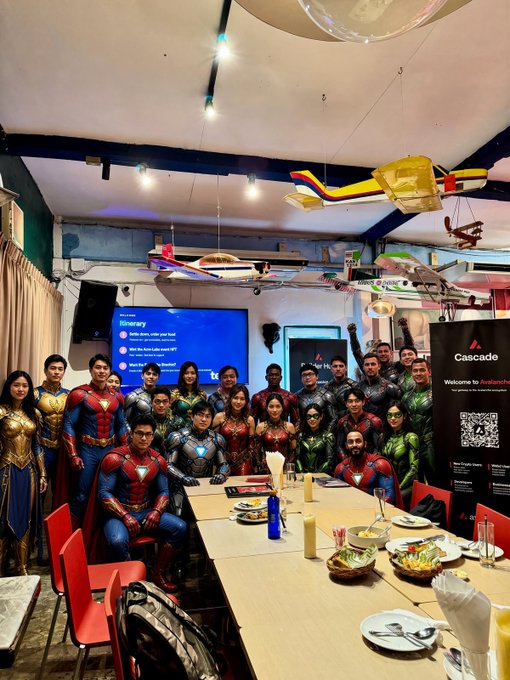 A group of people dressed in various superhero costumes, including Superman, Iron Man, Wonder Woman, and Spider-Man, gathered around a long table with food and drinks in a restaurant setting.