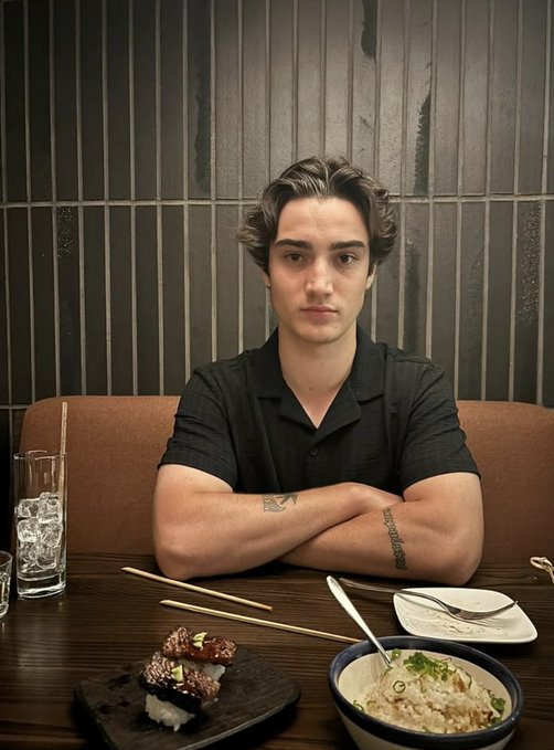 Portrait of a young man with wavy hair wearing a black shirt, seated at a restaurant table with his arms crossed in front of sushi rolls, chopsticks, and a bowl of food.
