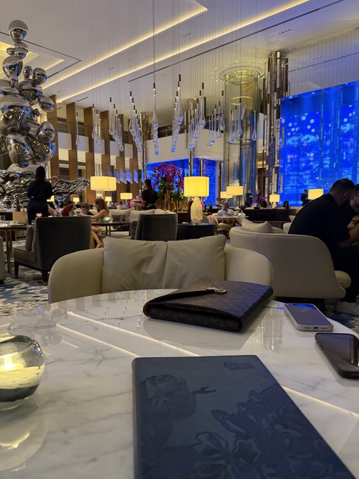 An elegant hotel lobby or lounge with modern decor, featuring a white marble table in the foreground with a dark Louis Vuitton-patterned wallet, an iPhone, and a blue menu, set against a backdrop of hanging lights and a blue illuminated wall.