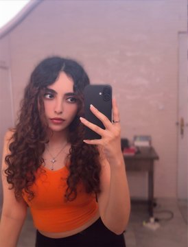 The image features a woman with curly hair wearing an orange top, taking a selfie using her cell phone. The background of the photo shows a white door and a brown wall, creating a contrast between the subject's appearance and the surroundings. The caption on the image reads "Good morning and happy Monday! The burden @TransientLabs 0.0033 eth per edition". This text is located at the bottom right corner of the photo.