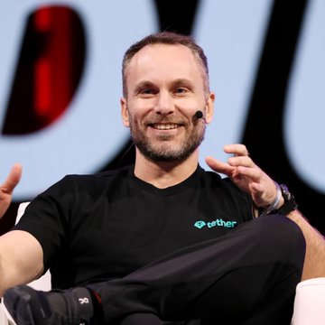 Tether CEO Paolo Ardoino smiles while speaking at a conference, wearing a black t-shirt branded with the Tether logo, as his company faces a lawsuit from cross-border payments firm Conduit over the freezing of $2.76 million in USDT during a Brazilian investigation.
