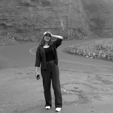 A black-and-white photo of a woman in a jumpsuit and sunglasses standing on a winding road with a rocky cliff in the background.