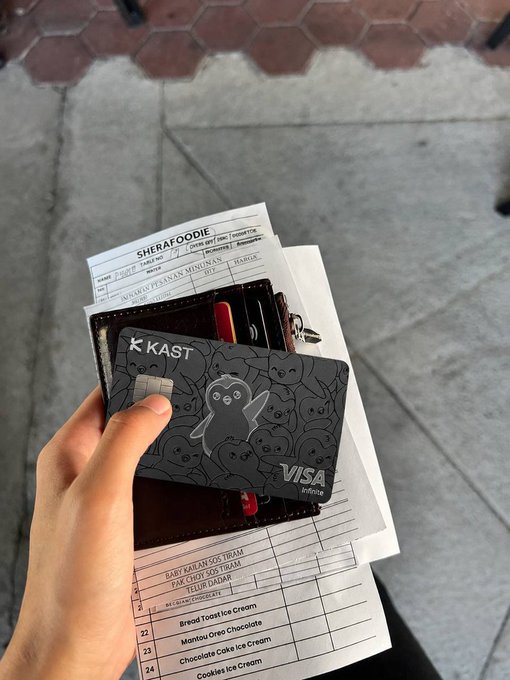 The image shows a person holding a black credit card with a cartoon character design, presumably from KAST, over some paperwork or bills. The person's hand is positioned above the papers, suggesting that they are about to use the card for payment. The scene also includes a wallet and a receipt, indicating that this might be related to a lunch paid service. The image focuses on the interaction between the credit card and the documents, highlighting how modern technology has made it easier than ever to manage financial transactions in our daily lives.