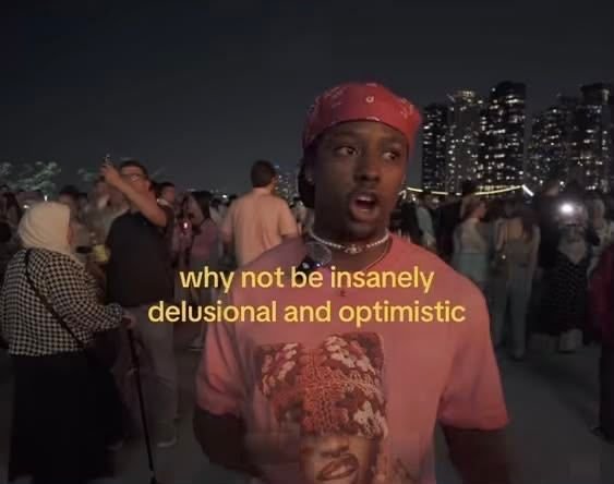 A TikTok video screenshot featuring a man in a red bandana and pink graphic t-shirt at an outdoor nighttime event, with the caption "why not be insanely delusional and optimistic."