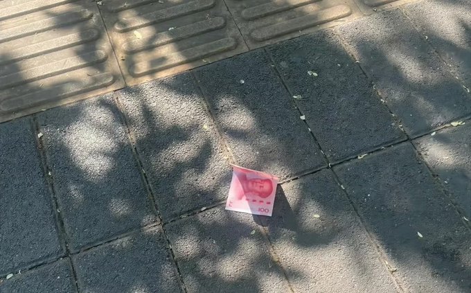 A pink 100 Turkish lira banknote lies crumpled on a paved sidewalk, partially shaded by tree shadows.