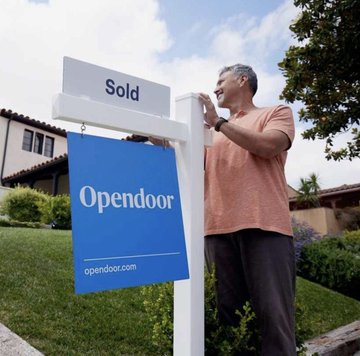 A man stands beside a white real estate sign reading "Sold" and a blue "Opendoor" banner (opendoor.com) in front of a house, appearing to check his phone. The image illustrates the post's message of Bitcoin being used for tangible real estate transactions, turning dormant capital into productive on-chain investment in housing markets.