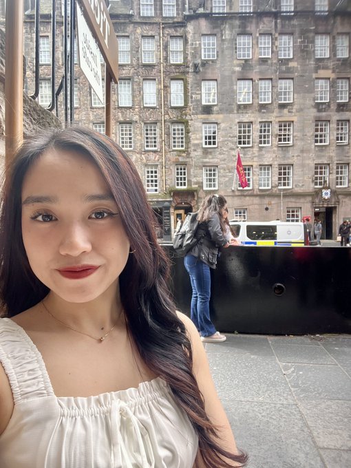 A selfie of a young woman with long dark hair wearing a white top and delicate necklace, taken outdoors with a historic stone building and a person with a backpack visible in the background.