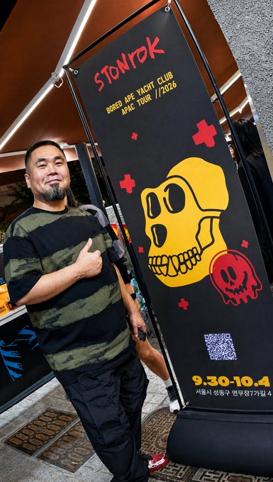 A man poses with a thumbs-up next to a black promotional banner for "STONROK Bored Ape Yacht Club ARIC Tour / 2026," featuring a yellow skull graphic and event dates "9.30-10.4." The banner ties into the BAYC x Stonrok pop-up collaboration mentioned in the post, showcasing one of its branded displays.