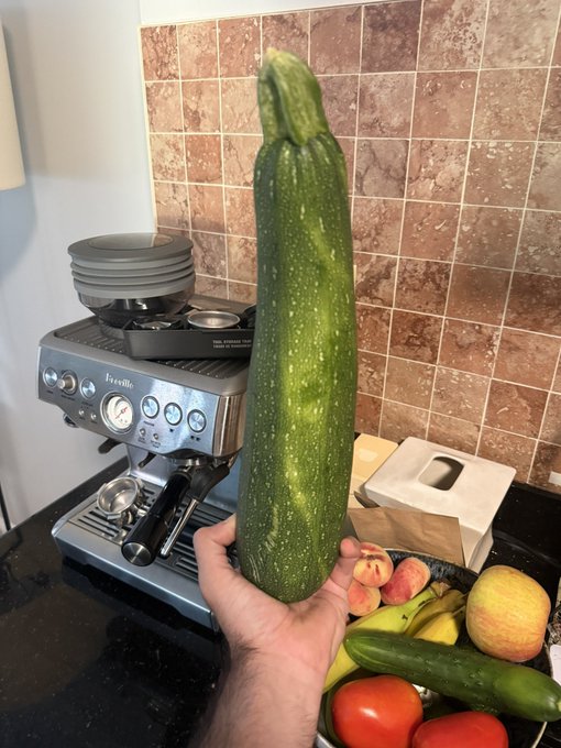 A hand holds up an oversized homegrown zucchini next to a "Breville" espresso machine, with a bowl of fresh fruit (apples, bananas, cucumber, tomato) on the counter. The post humorously compares the vegetable's towering green shape to Bitcoin's historic candlestick charts, using it as a tongue-in-cheek visual reminder of past crypto bull runs.