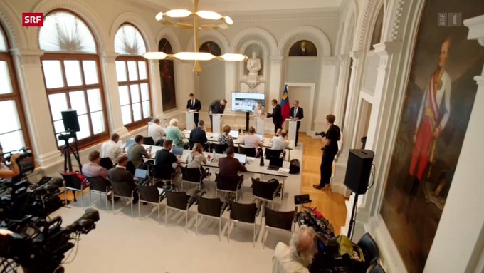 A press conference or media event in an ornate room with arched windows and classical portraits, featuring the SRF (Swiss Radio) logo, with people seated at a table and a speaker at a podium.