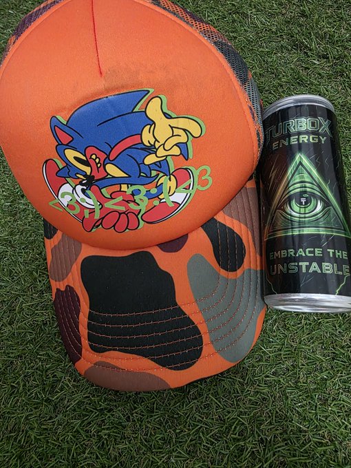 An orange camouflage cap featuring a Sonic the Hedgehog graphic rests on grass next to a black energy drink can labeled 'TURBO ENERGY' with an eye design and the slogan 'EMBRACE THE UNSTABLE'.