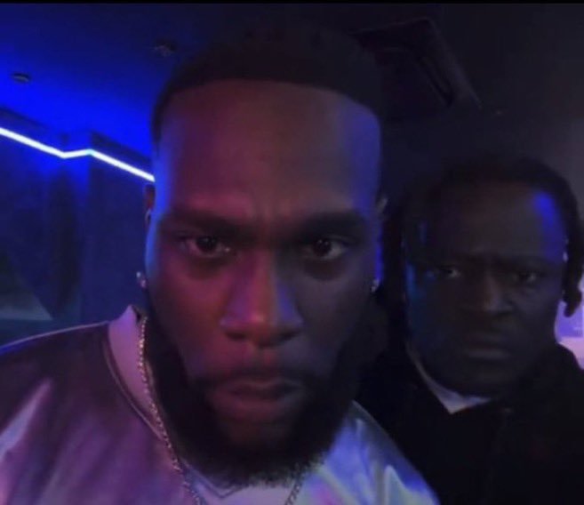 A close-up selfie of a man with a beard wearing a backward cap, looking at the camera in a dimly lit room with blue and purple lighting, while another man stands behind him.