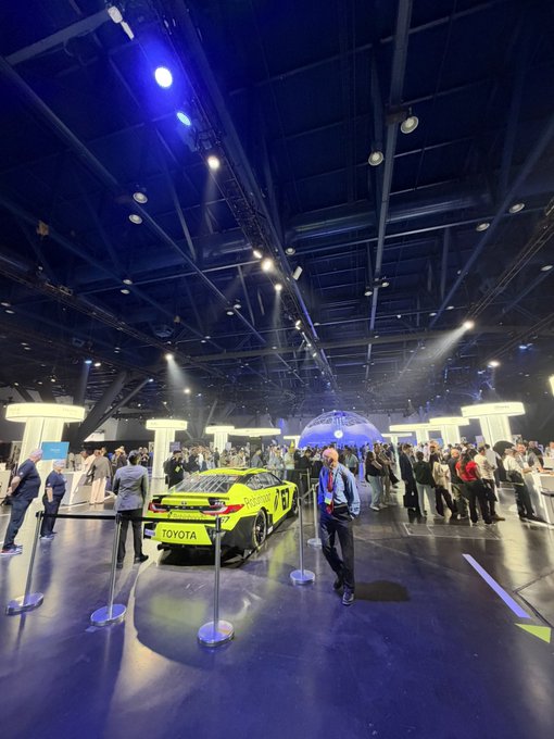 A bright yellow Toyota race car (#7) is on display at an indoor exhibition, surrounded by spectators under dramatic blue and white stage lighting. The "Hmm" caption suggests a contemplative reaction to seeing this striking racing vehicle on showcase.