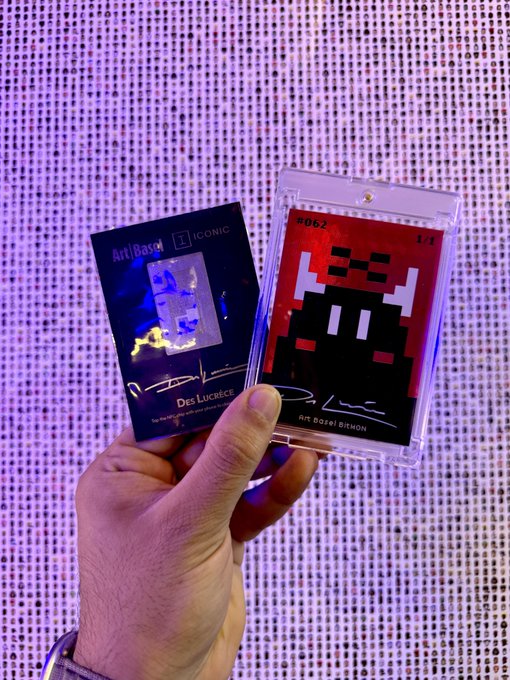 A hand holds up two collectible cards against a purple-lit background; the left card is titled 'ArtBoard ICONIC' featuring 'Des Lucrée,' and the right card is encased in a protective sleeve showing pixel art by 'ART BORIS BIHON.'