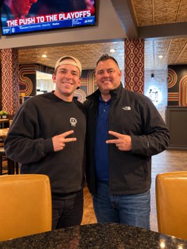 Two men pose for a photo in what appears to be a sportsbook or bar, with a TV screen above them displaying "THE PUSH TO THE PLAYOFFS".