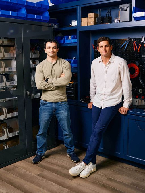 Two men pose in what appears to be an organized workshop or hardware lab with blue storage cabinets, plastic bins, and tools visible in the background. The caption suggests this is connected to Base Power Company (@basepowerco), featuring engineers or founders at their Austin HQ, likely promoting a hackathon event where participants can engage with Base's engineering team.