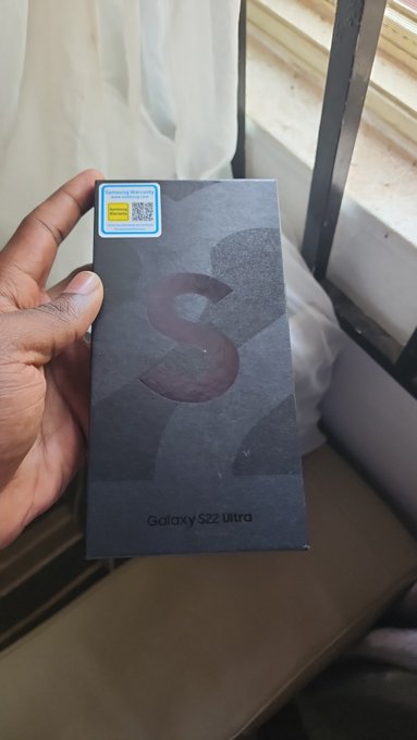 A hand holds a black Samsung Galaxy S22 Ultra smartphone box with a yellow authenticity sticker, set against a window with curtains.