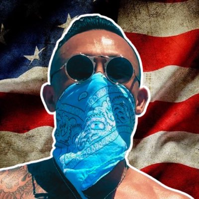 A man wearing sunglasses and a blue bandana face covering, with visible tattoos, set against a weathered American flag background.
