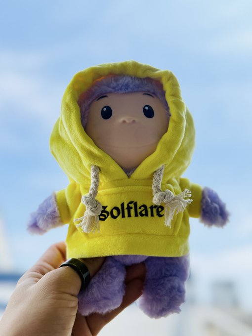 A purple plush doll wearing a yellow 'golfiare' hoodie, held up against a sky background.