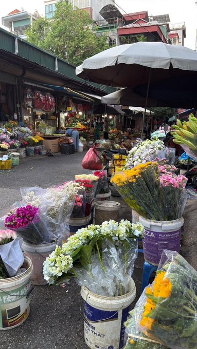 An outdoor flower market with numerous buckets filled with bouquets of flowers including sunflowers, roses, and white flowers wrapped in plastic, displayed under umbrellas and awnings.