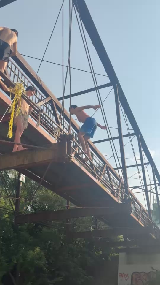 A group of young people, possibly skateboarders, are perched precariously on the railing of a rusting metal bridge under a clear sky, with one mid-leap off the edge—evoking the reckless energy of "bailing" a trick, which ties directly to the post's question about stopping from bailing flips.