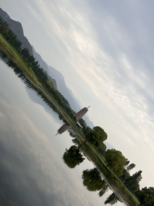 A serene landscape photograph taken at a tilt shows a calm lake reflecting trees, a pagoda-style building, and distant mountains under a cloudy sky.