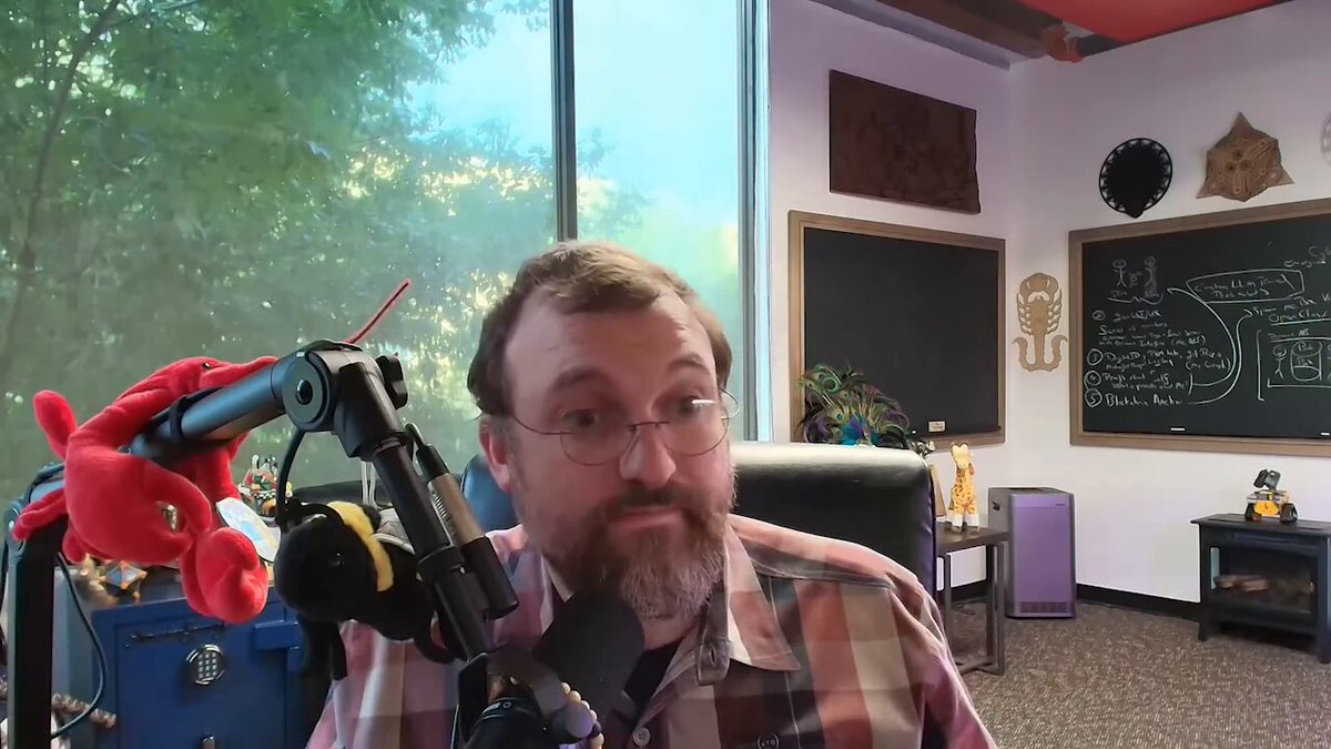 The image features a man wearing glasses sitting behind a microphone, likely participating in an interview or podcast recording session. The man has a beard and is dressed in a plaid shirt. In front of him, there are two stuffed animals - one red and one yellow. A window can be seen in the background, providing natural light to the room. The image also contains text that reads "UPDATE: #Cardano $ADA Founder Charles Hoskinson says Cardano has very special stuff. We predicted all the DeFi hacks, which is why we embraced formal methods. All coding is moving to specifications, or in our case, formal specifications."