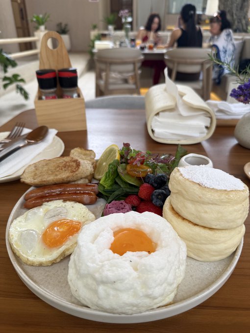A cloud-like soufflé pancake sits center stage on a breakfast plate, surrounded by fluffy eggs, sausages, fresh berries, and fluffy Japanese-style pancakes dusted with powdered sugar. The image captures a leisurely brunch setting that perfectly embodies a relaxed "good morning" vibe.